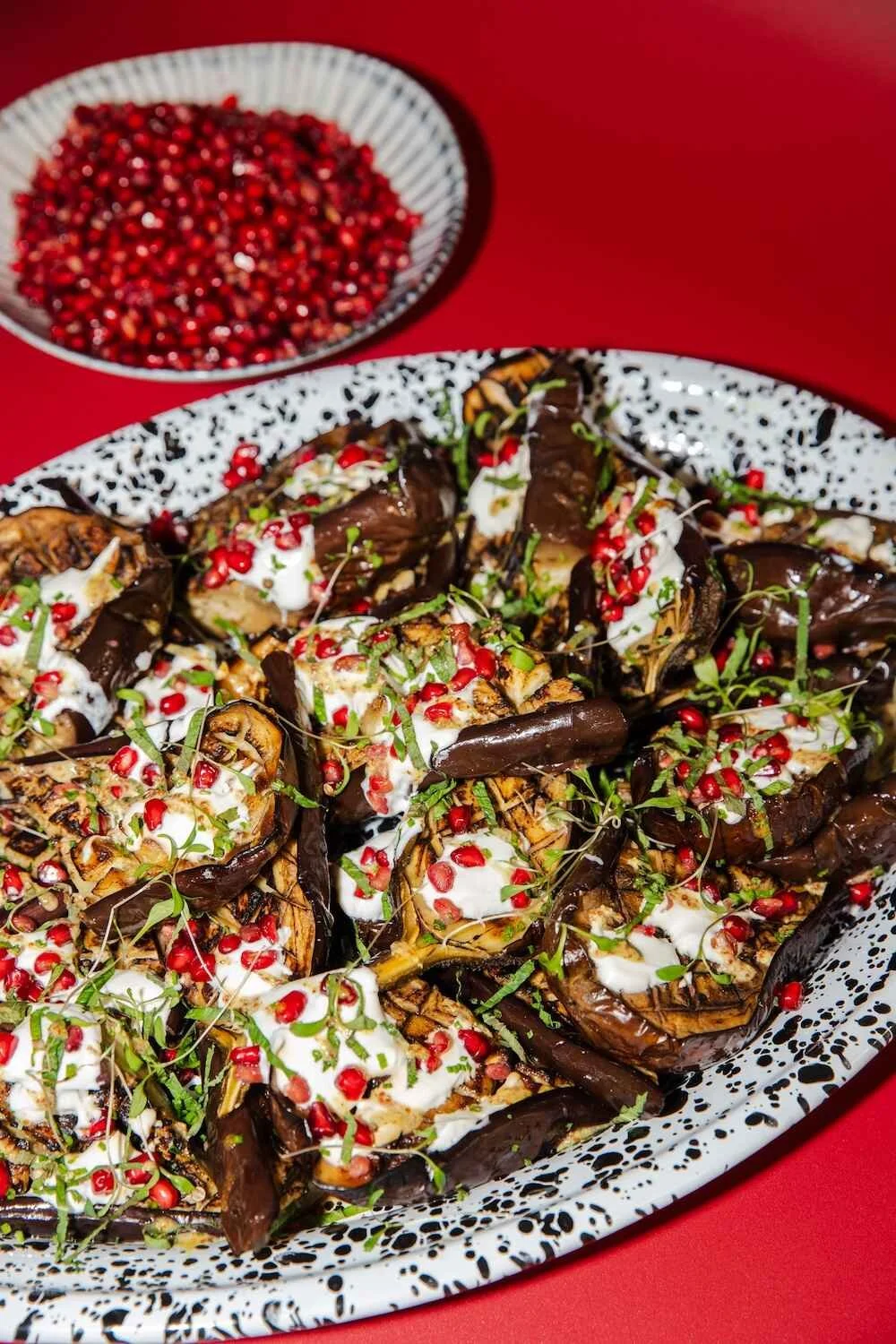 Dish of roasted eggplant topped with yogurt, pomegranate seeds, and microgreens on a speckled oval platter, with a bowl of pomegranate seeds in the background, set on a red surface.