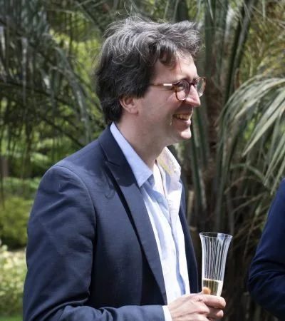 A man with glasses and dark hair, wearing a navy blazer and white shirt, smiling and holding a glass of wine outdoors in a lush garden setting.