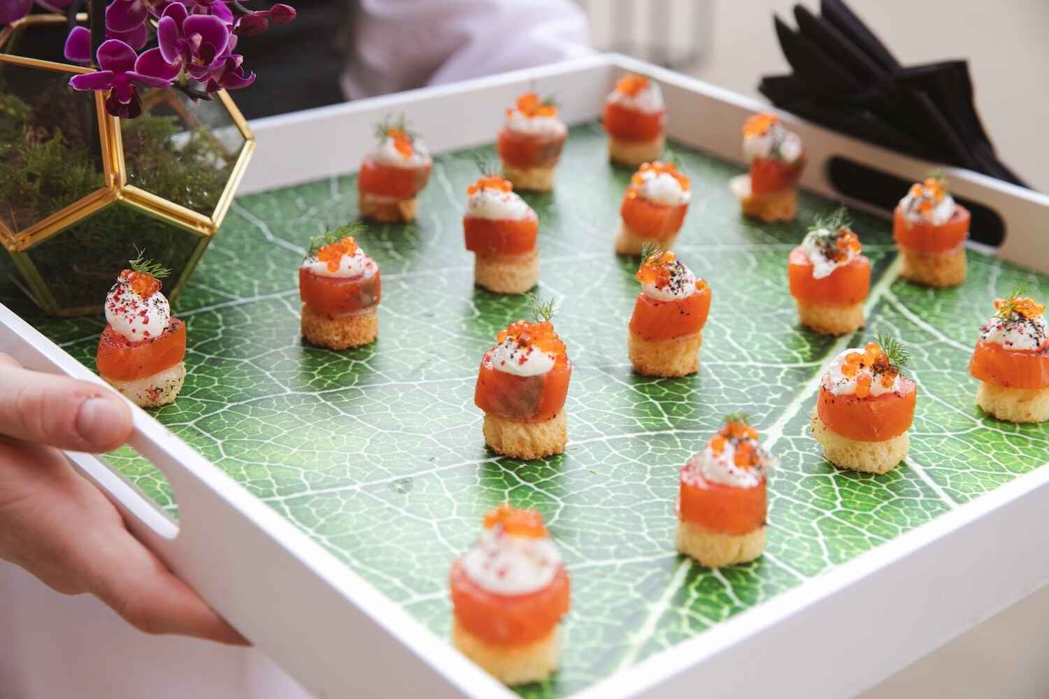 Tray of small appetizer bites with salmon, cream cheese, and herbs, decorated with orange fish roe, on a leaf-patterned paper, in a flower-shaped glass terrarium.