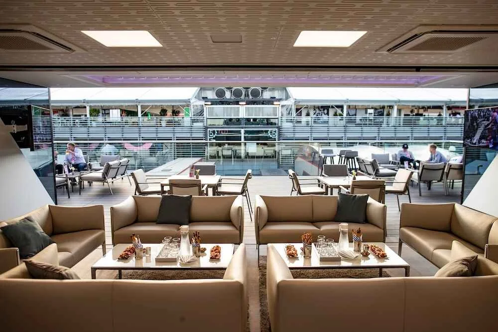 Modern airline lounge with beige sofas, black pillows, and a coffee table in the foreground, outdoor seating with tables and chairs beyond, and a glass wall separating the interior from the outdoor area.