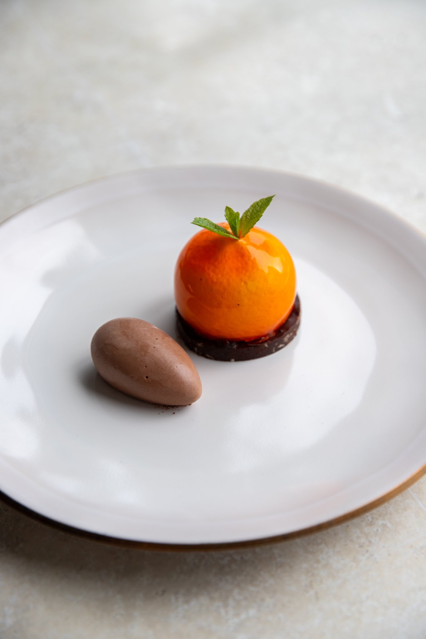 Dessert featuring an orange mousse with a mint leaf on top, served on a white plate with a chocolate base and a chocolate quenelle nearby.