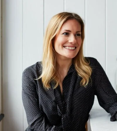A smiling woman with blonde hair, wearing a black blouse with white polka dots, sitting against a white paneled wall.