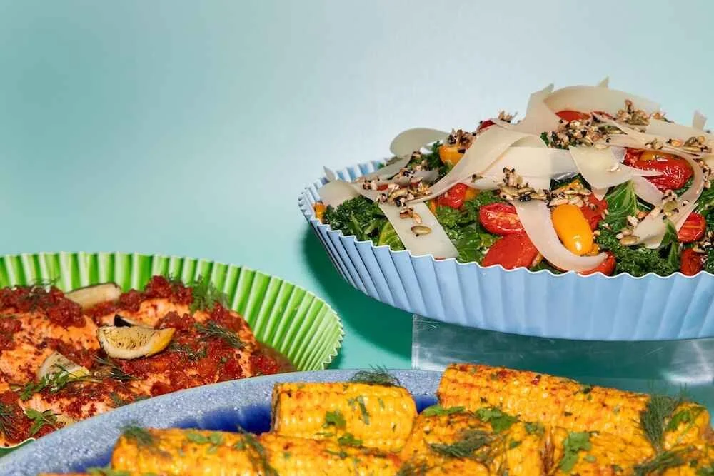 A colorful spread of three different dishes on pastel-colored plates and bowls, including a vegetable salad, seasoned grilled chicken, and corn with herbs.