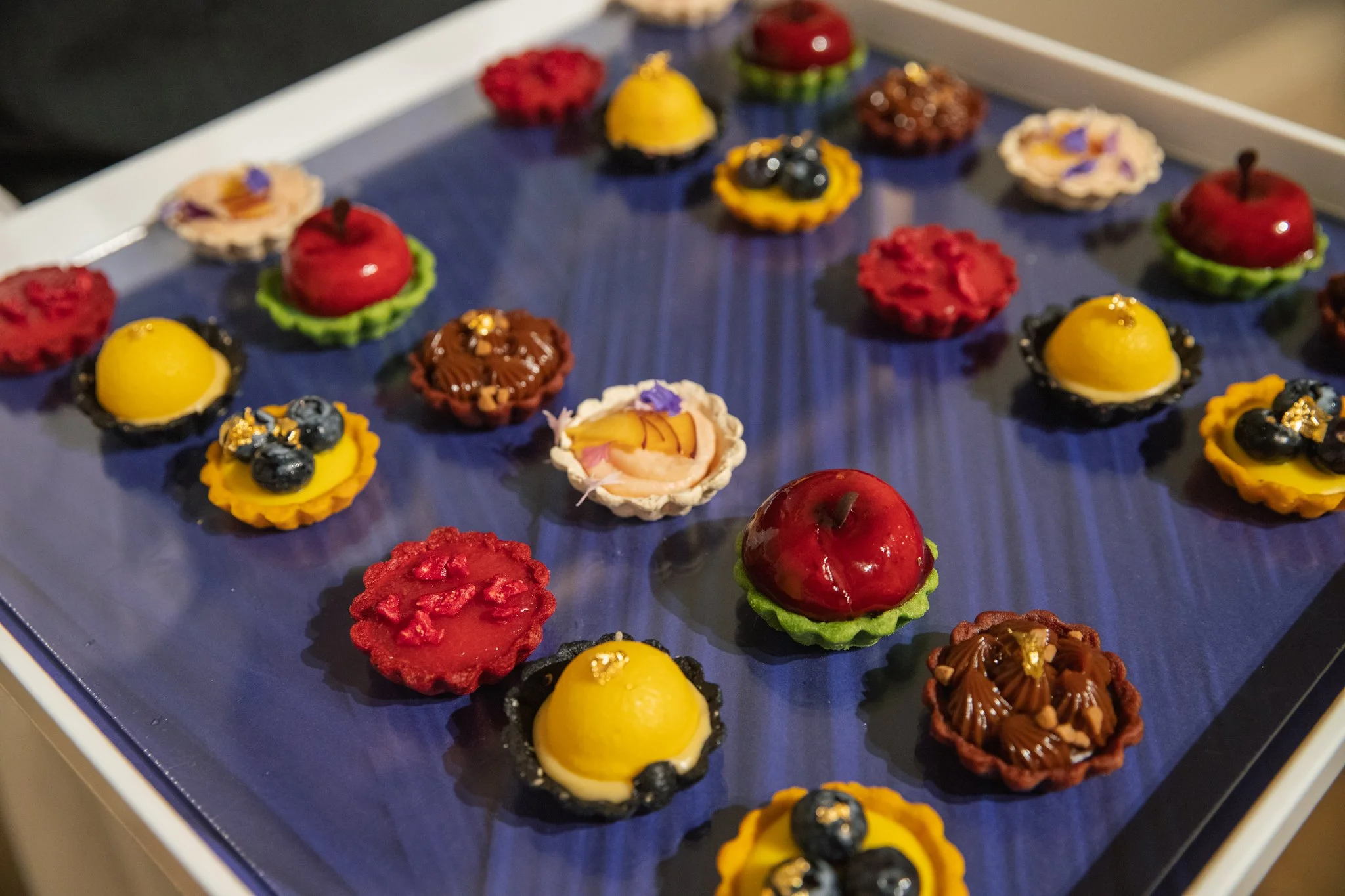 Assorted mini desserts on a blue tray, including fruit tarts, chocolate cups, and mousse cups decorated with berries, edible gold, and colorful toppings.