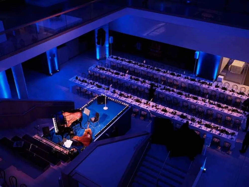 Empty venue with long dining tables with chairs, a small stage with a grand piano and a microphone, and blue lighting creating a concert or event space atmosphere.