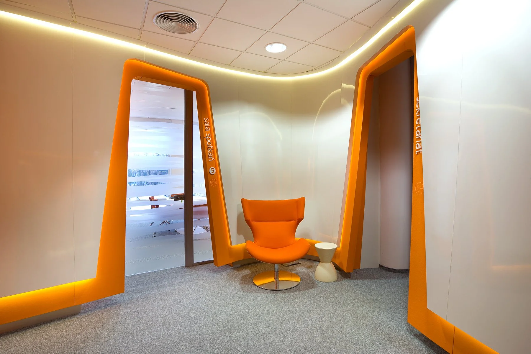 An orange modern chair and small white side table in a minimalist office space with curved white walls and orange accents, with glass door revealing a conference room.