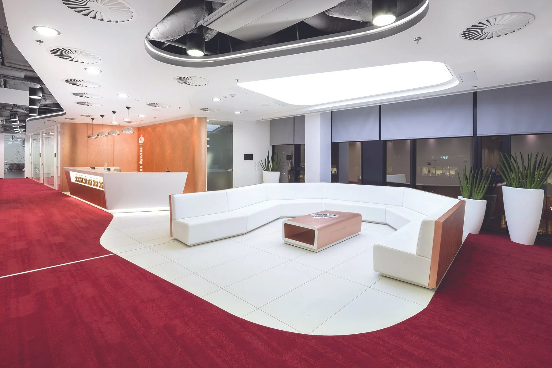Modern office lobby with white curved seating, a wooden coffee table, potted plants, a reception desk in the background, large windows with roller blinds, and red carpet flooring.