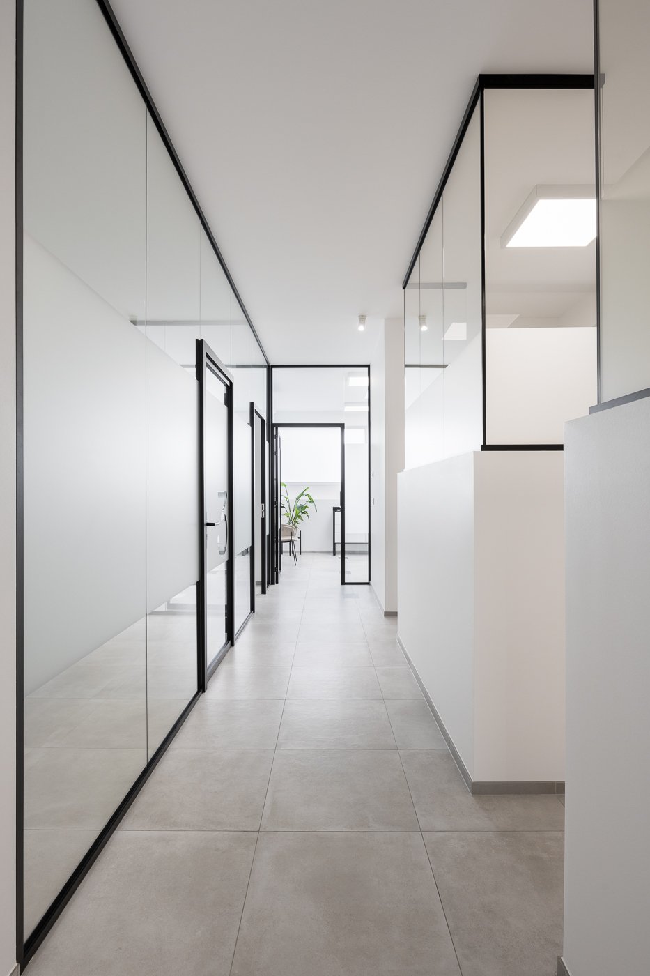Minimalist office corridor with glass walls, white walls, and gray tiled flooring, leading to a bright room with a plant and chairs.