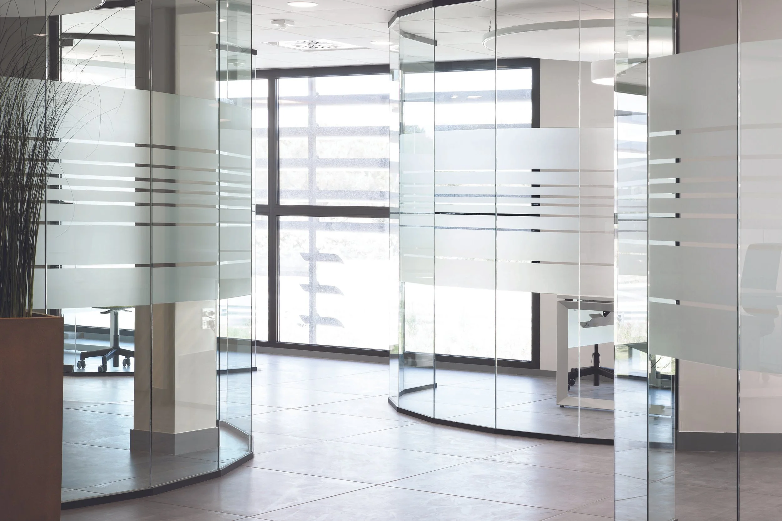 Modern office space with glass walls, a large window, a rolling chair, and a white desk.
