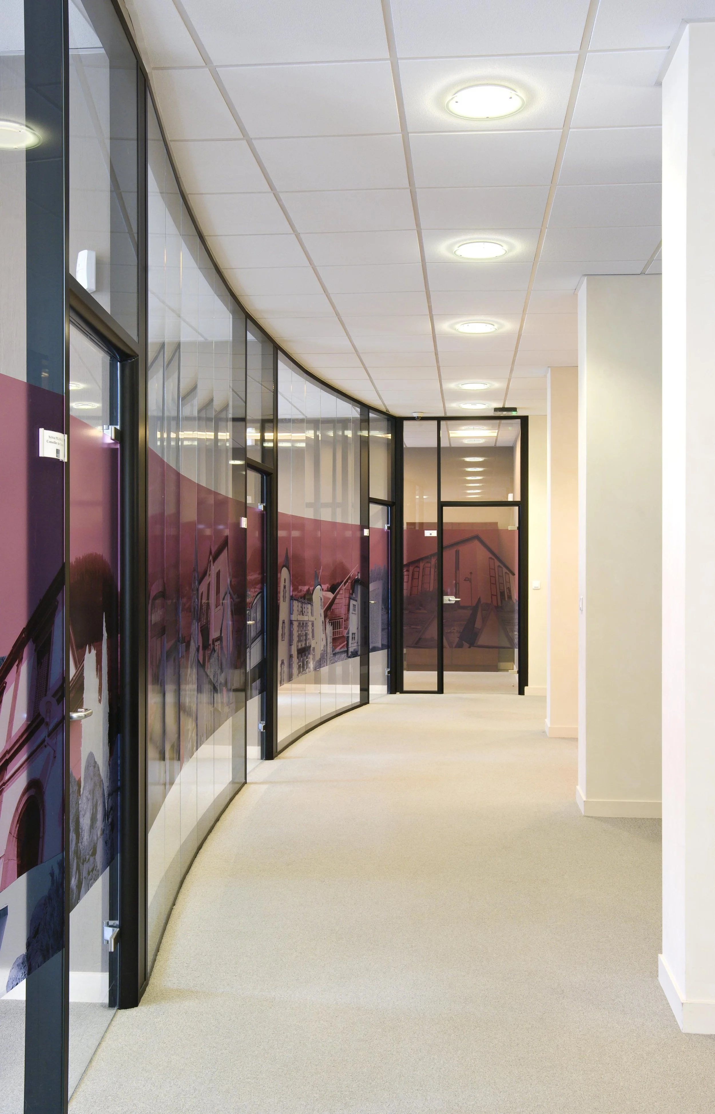 Interior of an office corridor with a curved wall of glass partitions featuring a mural of buildings and cityscape, cream carpet flooring, suspended ceiling with round light fixtures.