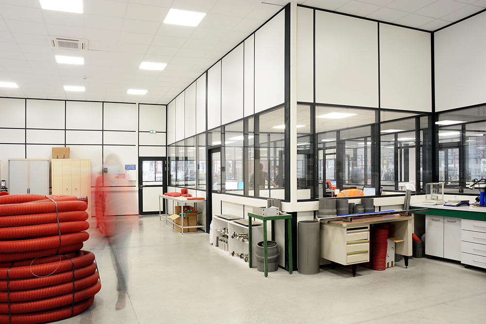 Industrial workspace with coiled red hoses, workstations, cabinets, and a glass partitioned room, in a well-lit environment.