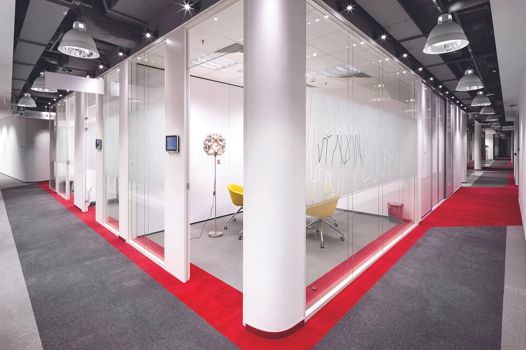Modern office conference room enclosed by glass walls with a white interior, yellow chairs, and a unique spherical lamp, in a hallway with gray and red carpet.