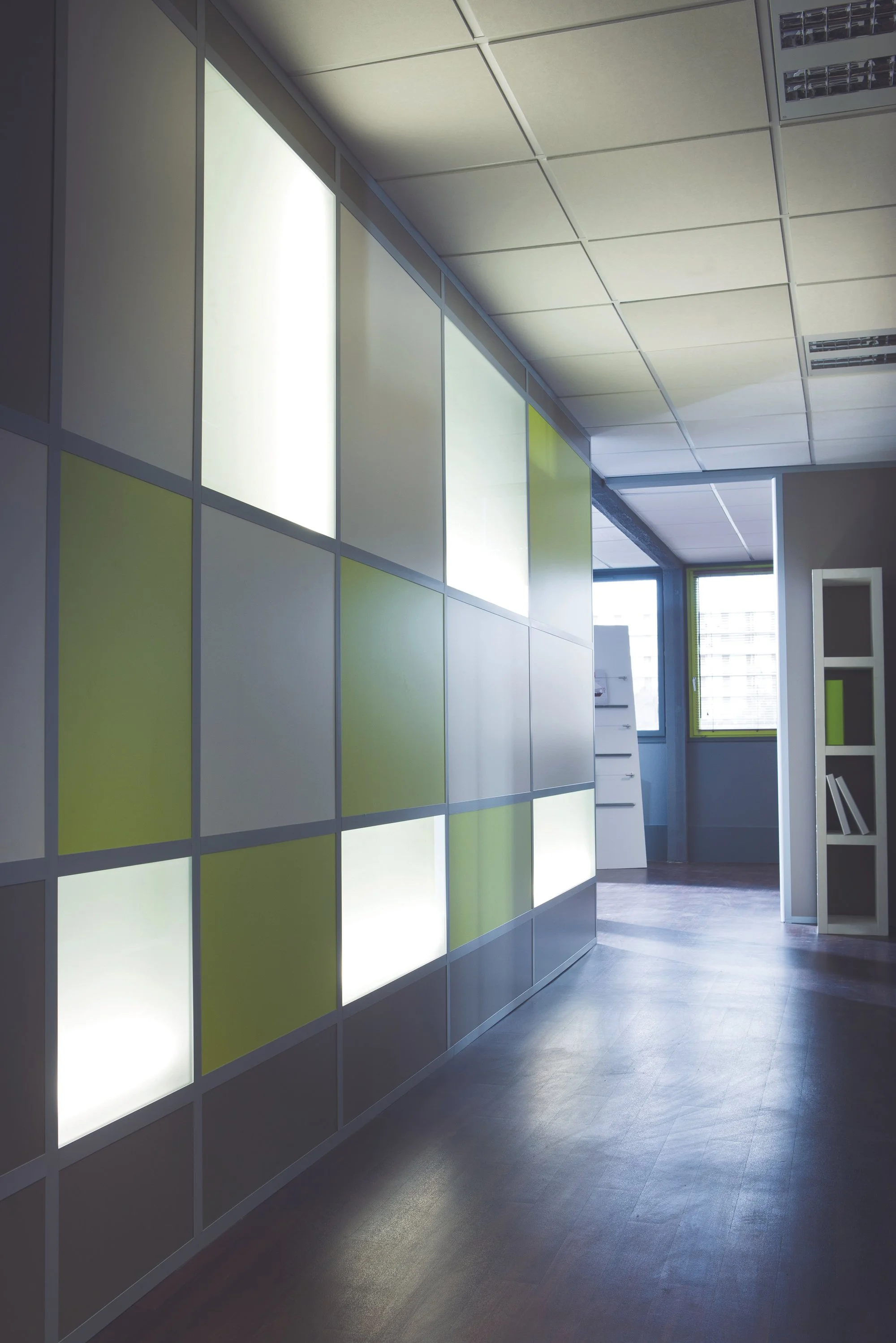 An interior office space with a modern design, featuring a partition wall made of illuminated panels in white and light green. There is a window at the back with sunlight coming through, and a white bookshelf with a few notebooks or files on the right side.