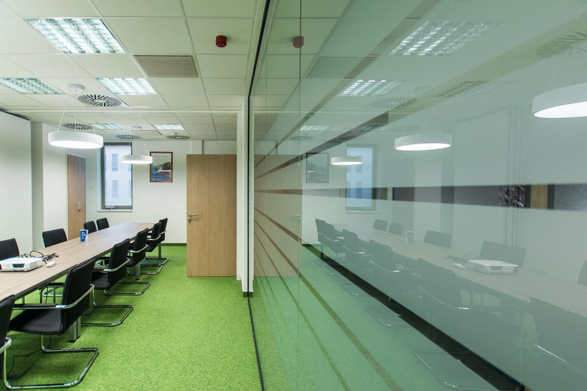 Empty conference room with black chairs around a rectangular table, green carpet, white walls, large window, framed picture, closed wooden door, ceiling lights, and a glass wall reflecting the room.