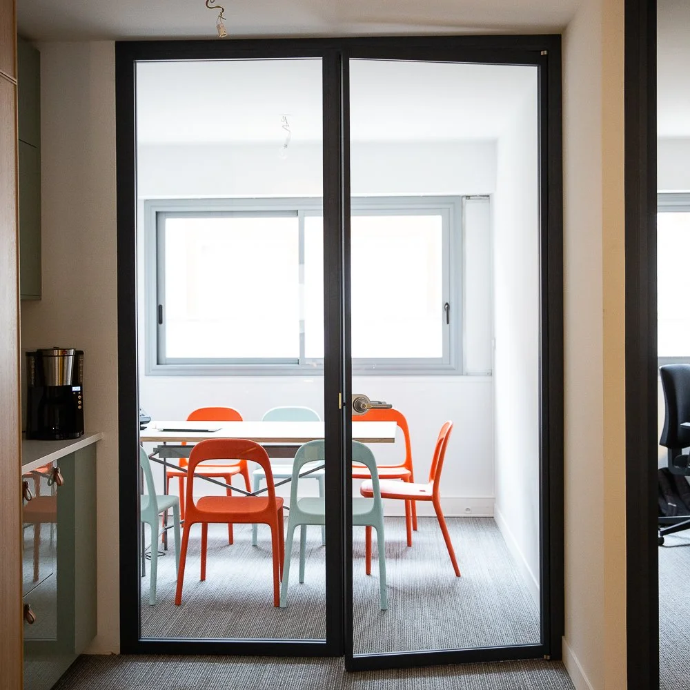 Modern office conference room with a glass sliding door, colorful chairs, large window, and natural light.