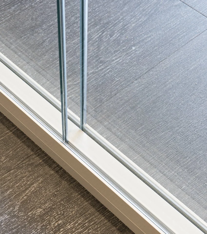 Close-up of a sliding glass door with a metal frame, showing the edge of the door and glass panels with a textured wooden floor outside.