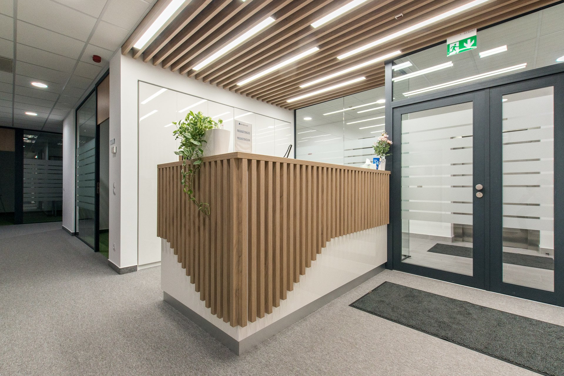 Modern office reception area with a wooden reception desk, glass doors, and fluorescent lighting.