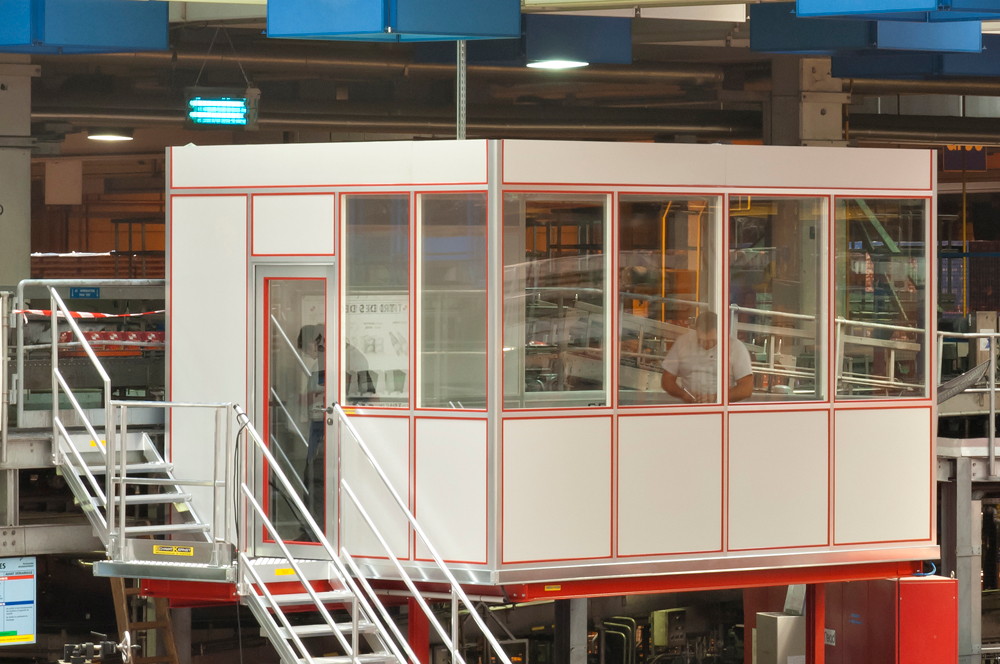 An industrial control room enclosed in glass with metal stairs leading up to it, situated in a warehouse or factory environment.