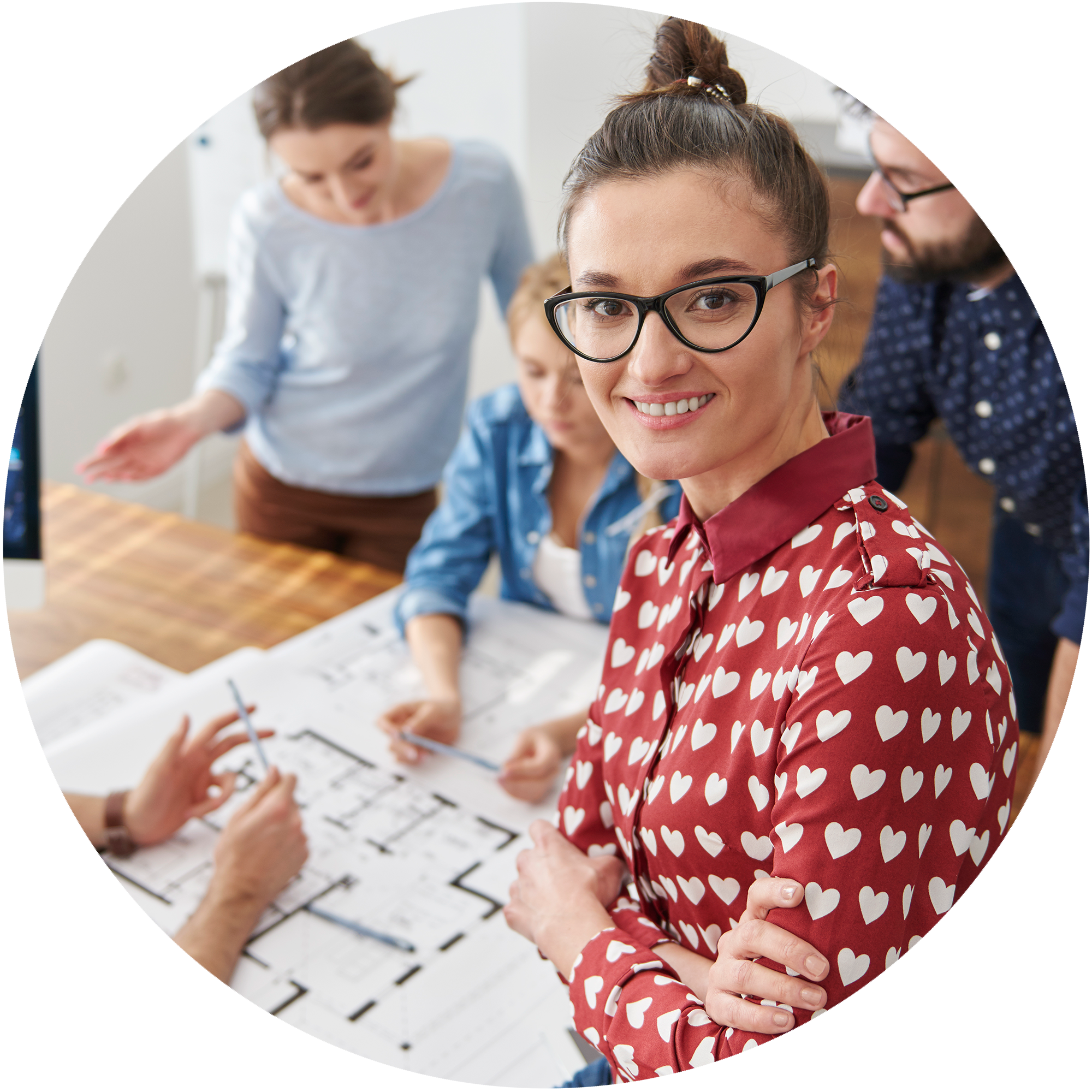 A woman with glasses and a red shirt with white hearts smiling at the camera, surrounded by colleagues working on architectural plans.