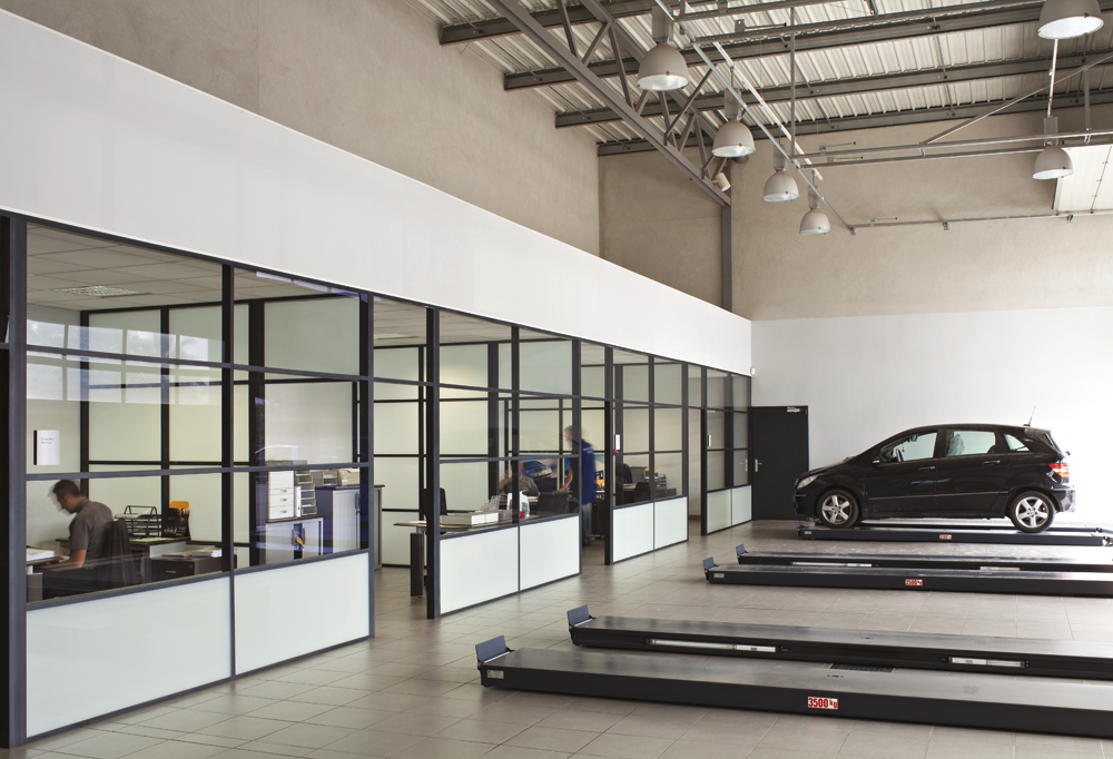 Car service garage with a black car on a lift and an office area with people working inside.