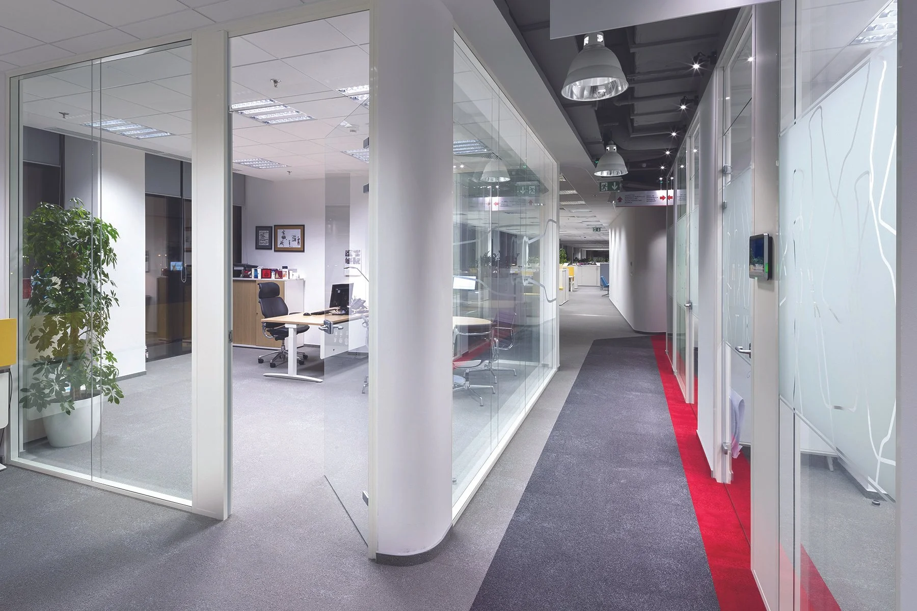 Empty office hallway with glass-wall conference rooms on the left and a corridor with a gray and red carpet on the right.