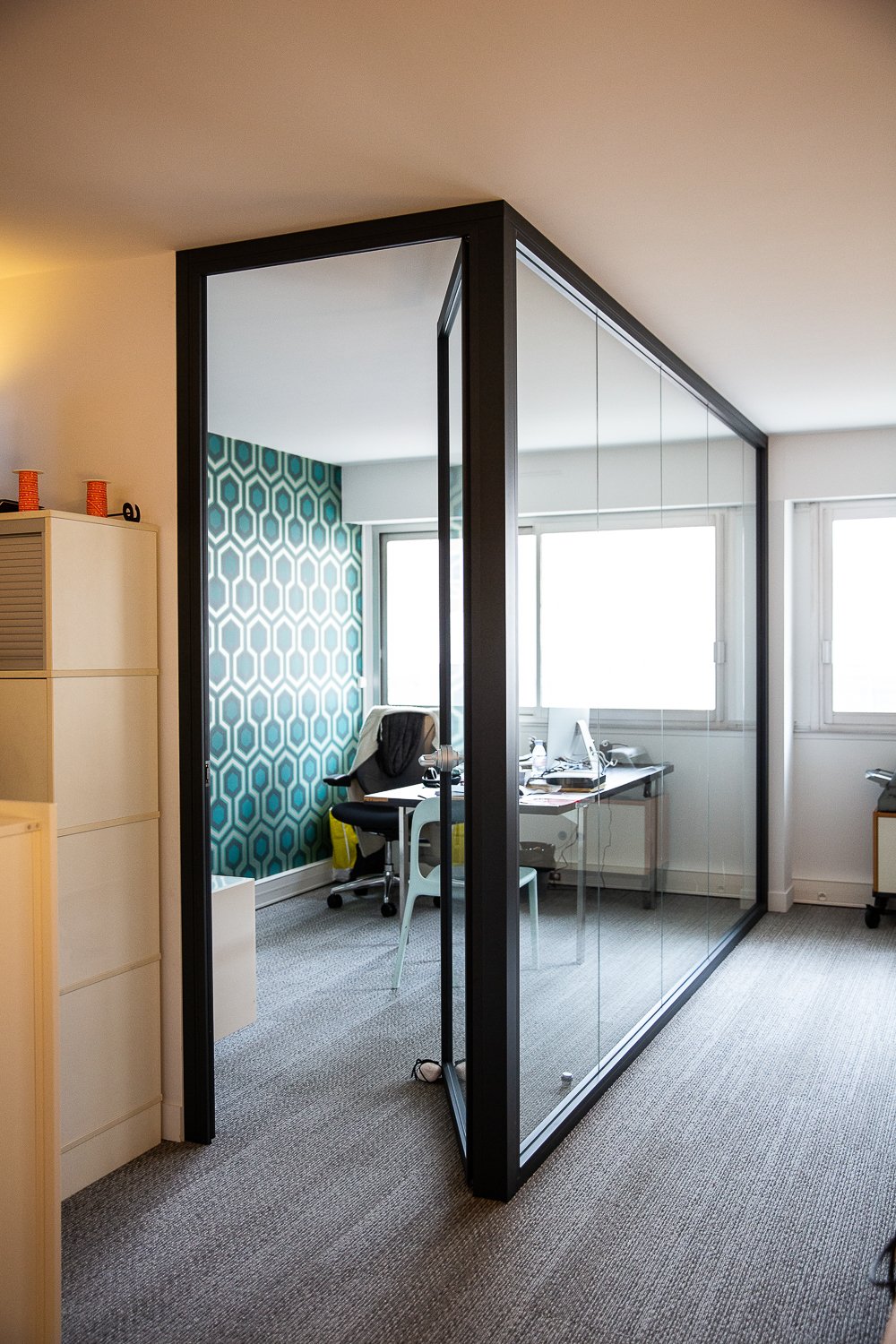 Office space with a glass-walled room and patterned accent wall, desk with computer, office chair, and window.