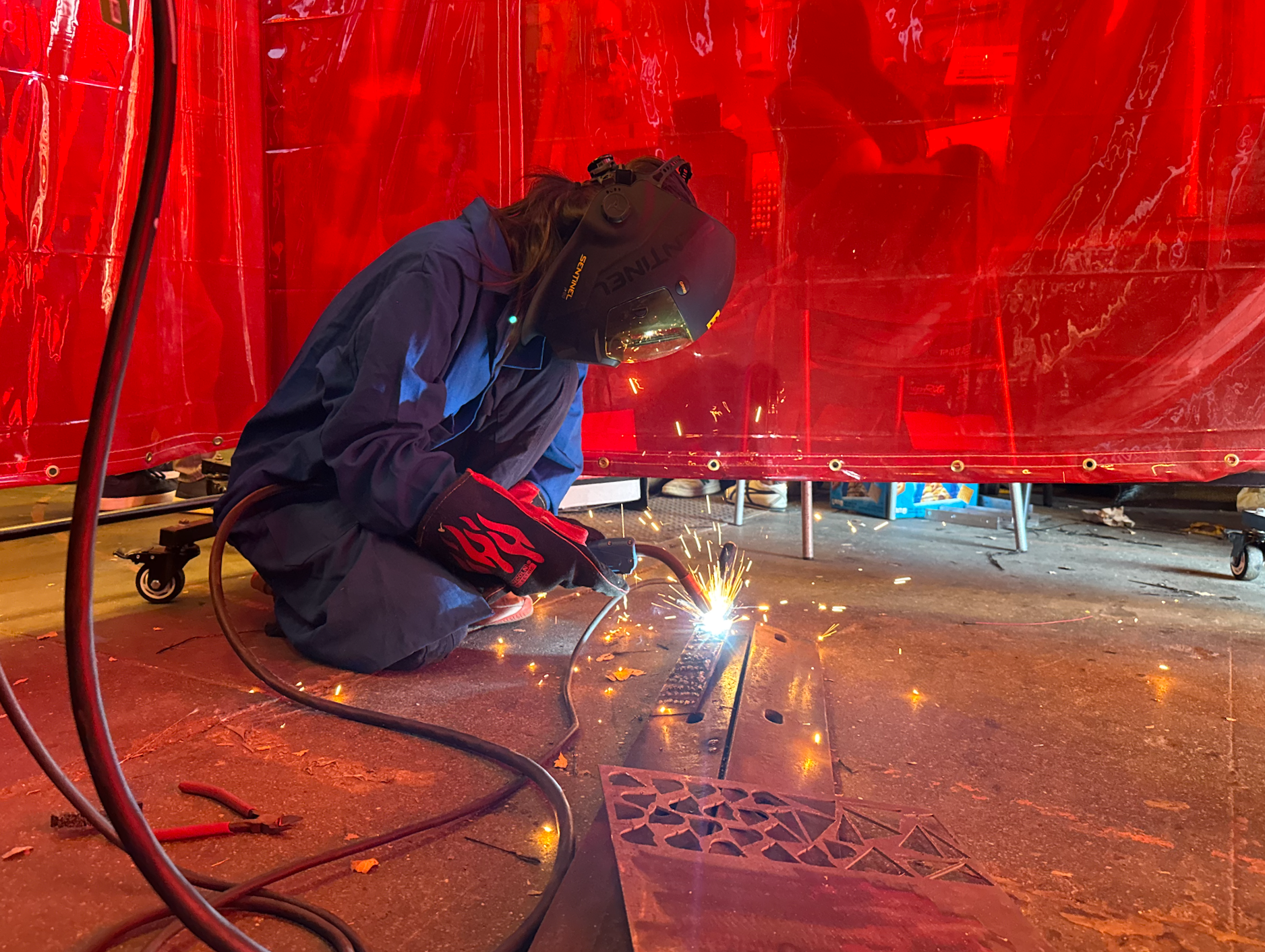 A person welding metal in a workshop, wearing a dark protective helmet and gloves, with sparks flying as they work on a metal piece.