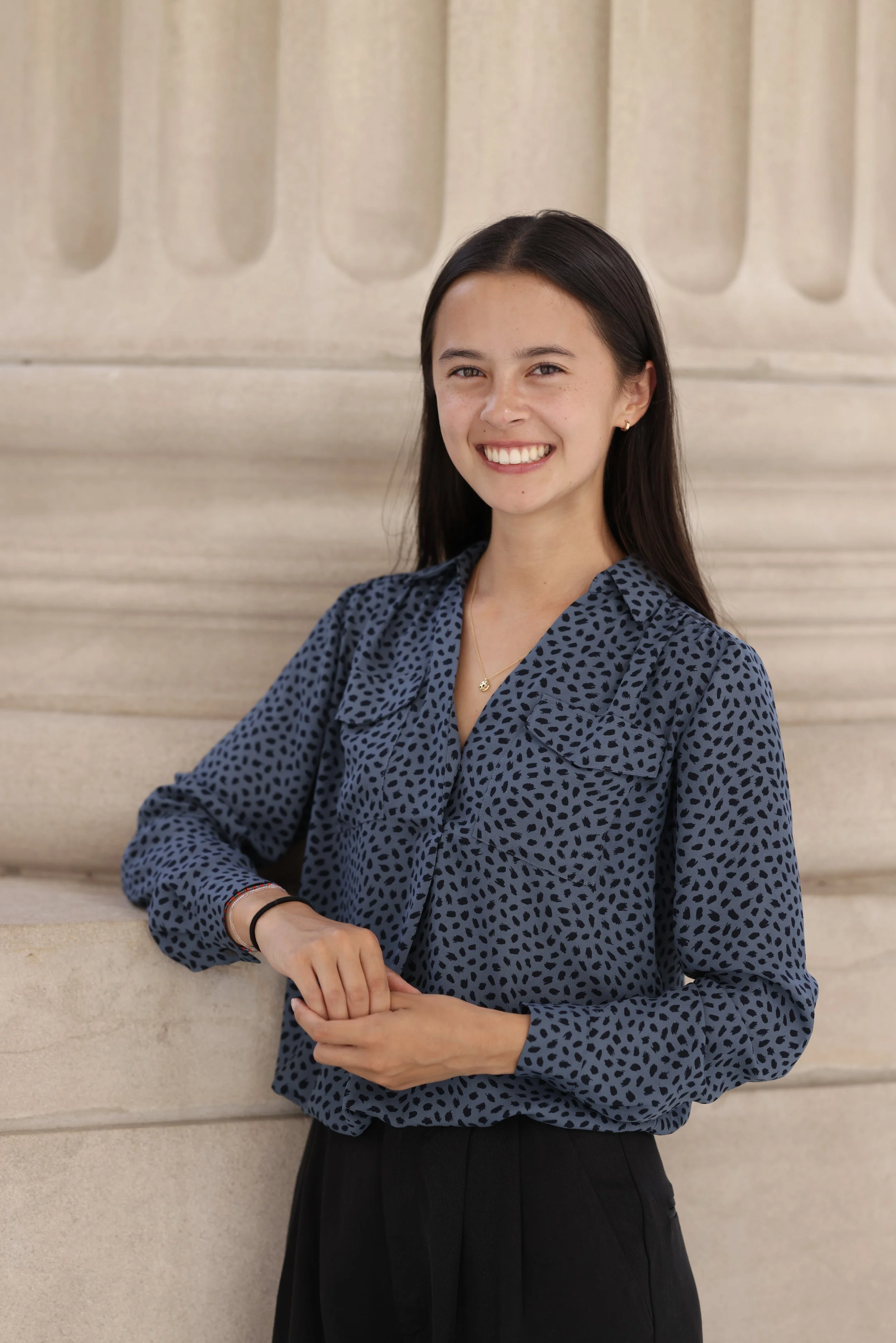 A young woman with long brown hair and a smile, standing with her arms crossed in front of a beige stone background, wearing a blue patterned blouse.
