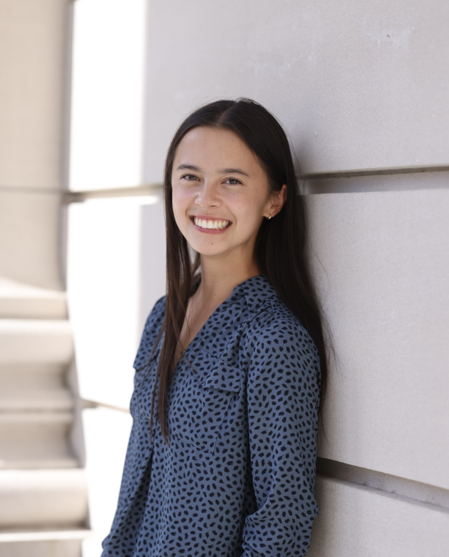 Young woman with long dark hair smiling, leaning against a concrete wall, wearing a blue patterned blouse, outdoors during daytime.