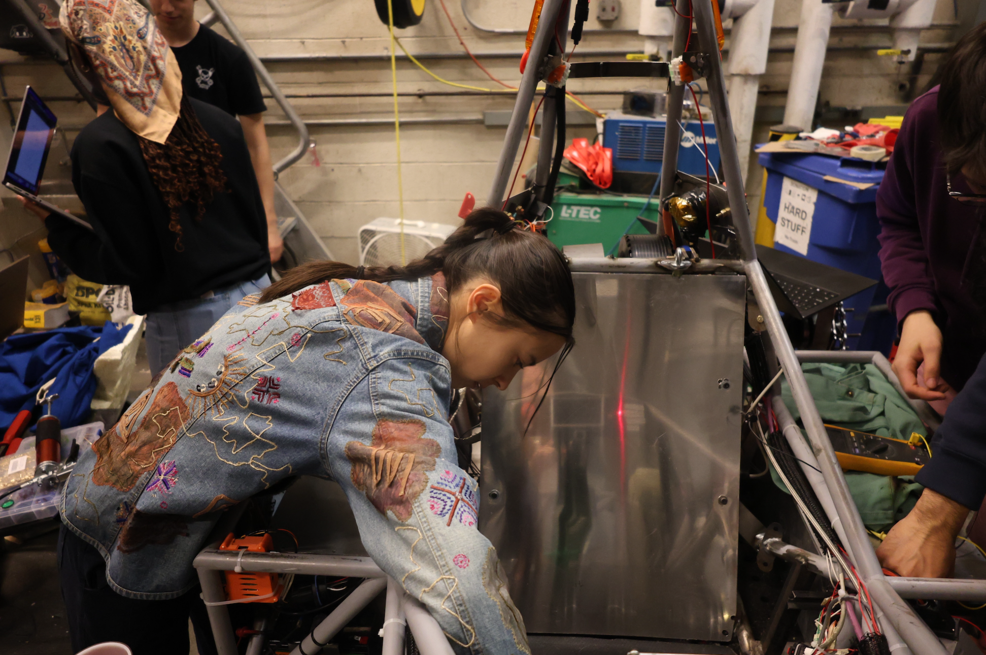 A woman in a denim jacket working on a metal frame structure with electronic wires inside a workshop. Other people are nearby, with tools and equipment visible.