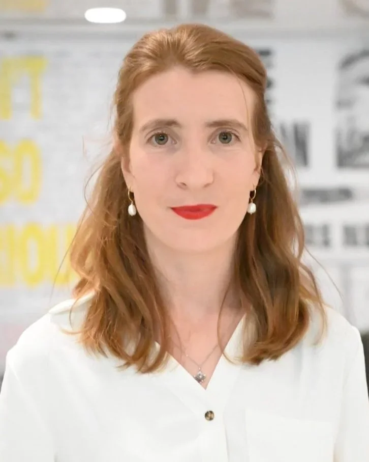A woman with red hair, wearing earrings and a white blouse, smiling at the camera in an indoor setting.
