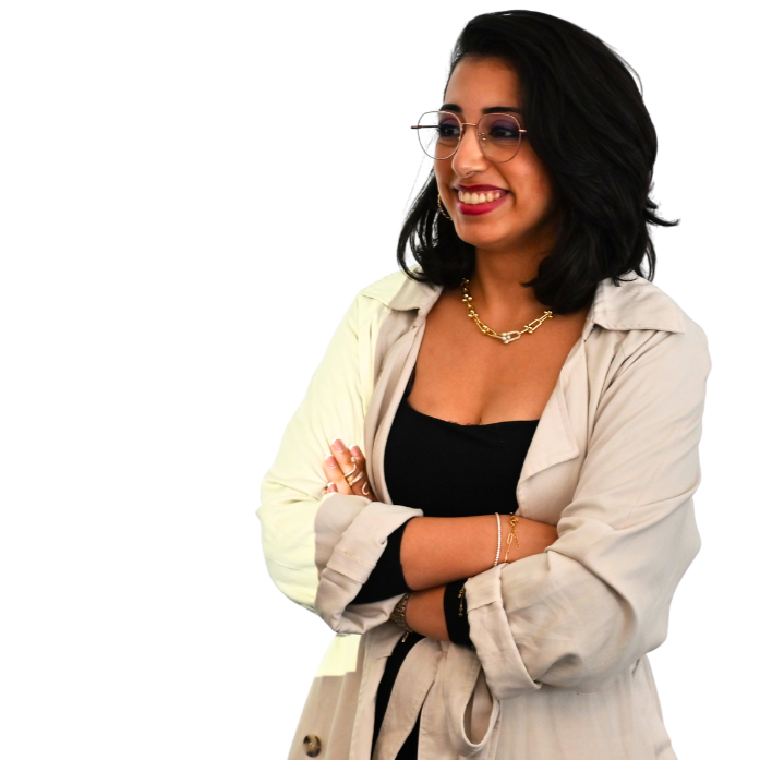 A smiling woman with dark hair wearing glasses, a gold chain necklace, and a beige trench coat, standing with her arms crossed.