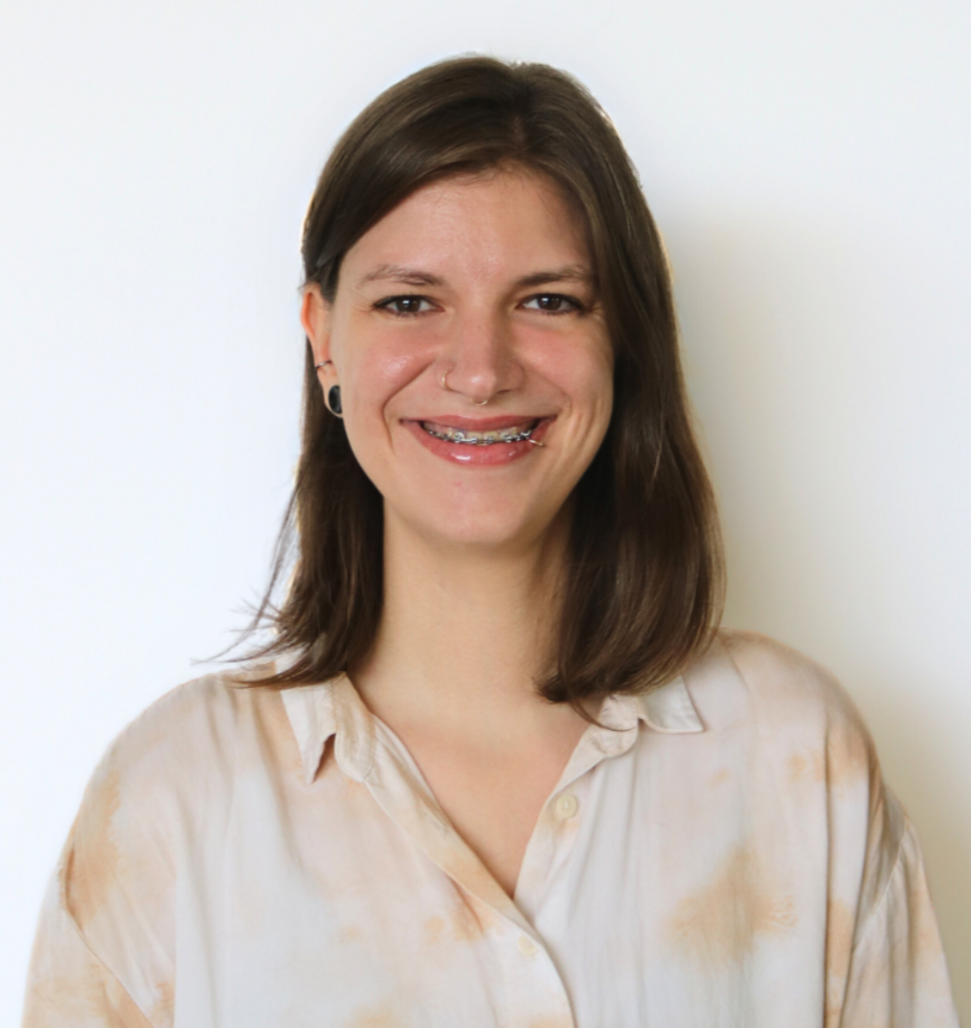 A woman with shoulder-length brown hair, wearing a light-colored shirt with a subtle pattern, smiling at the camera against a plain white wall background.