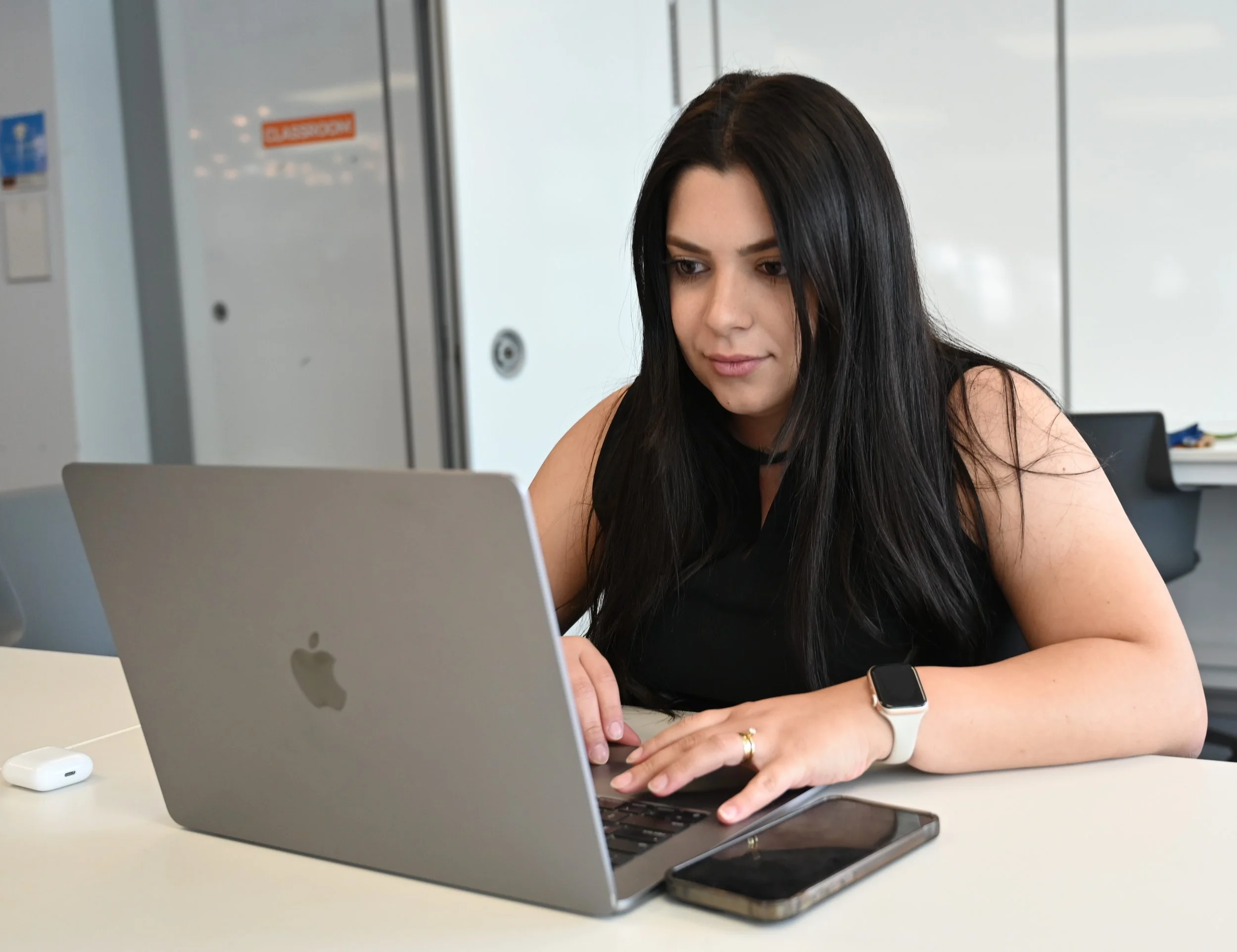 A woman with long black hair working on a silver MacBook at a white table in an office setting. She is wearing a black sleeveless top, a smartwatch, and a ring, with a smartphone and wireless earbuds on the table.