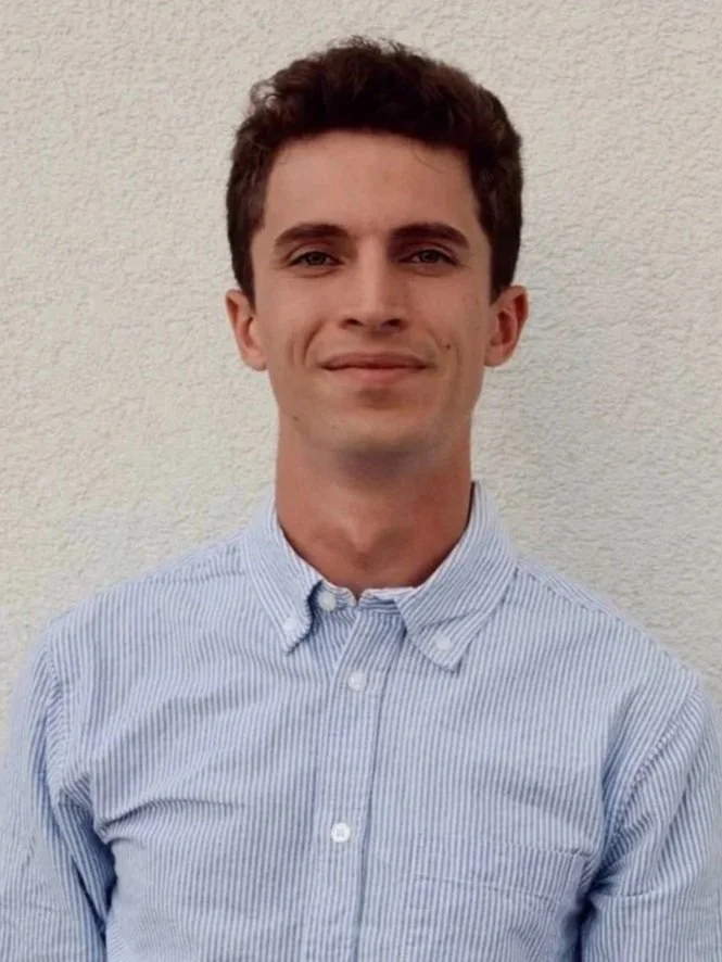 A young man with short dark hair, wearing a light blue striped button-up shirt, standing in front of a plain off-white wall, smiling lightly.