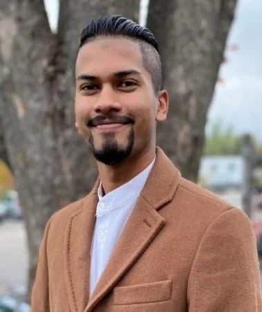 A man with a beard and mustache, wearing a brown coat and white shirt, smiling outdoors with trees in the background.