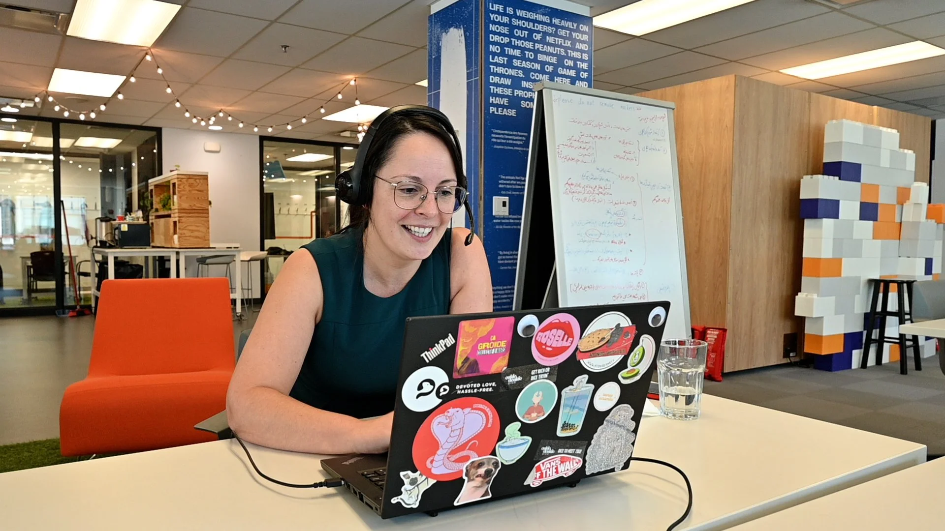 A woman wearing glasses and a headset sits at a table in a modern workspace, smiling at her laptop decorated with stickers, with a glass of water beside her.