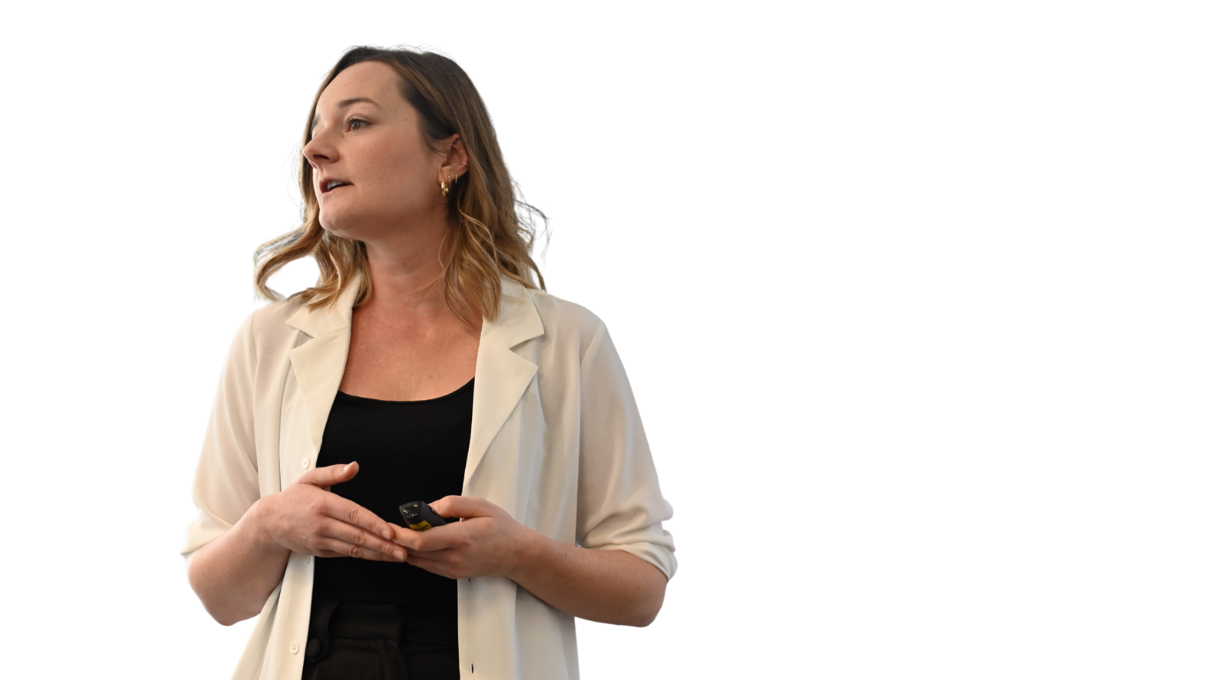 A woman with shoulder-length light brown hair wearing a cream-colored blazer over a black top, holding a wireless device, possibly a remote or a clicker, while speaking or presenting.