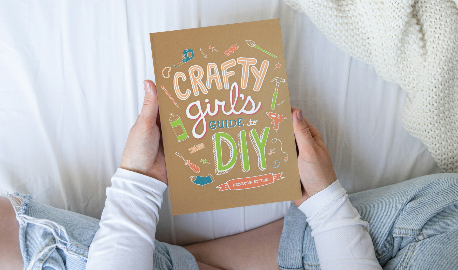 Person holding a beige-colored book titled 'Crafty Girls Guide to DIY, Bedroom Edition' with colorful illustrations of tools and craft supplies.