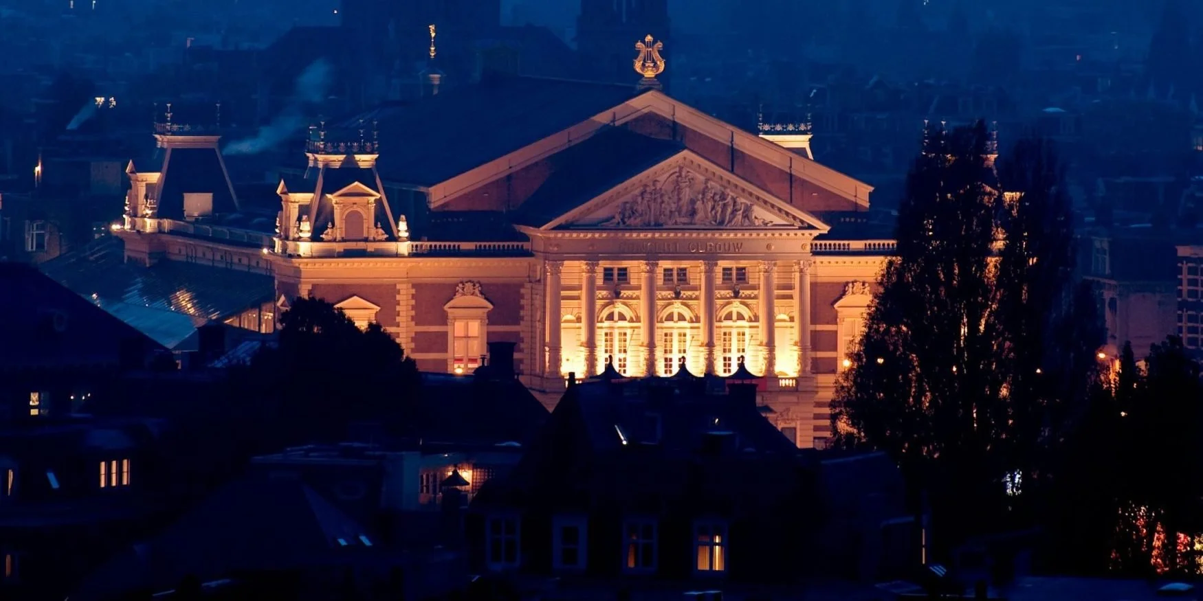 Illuminated historic building with classical architecture at night, surrounded by dark trees and rooftops.