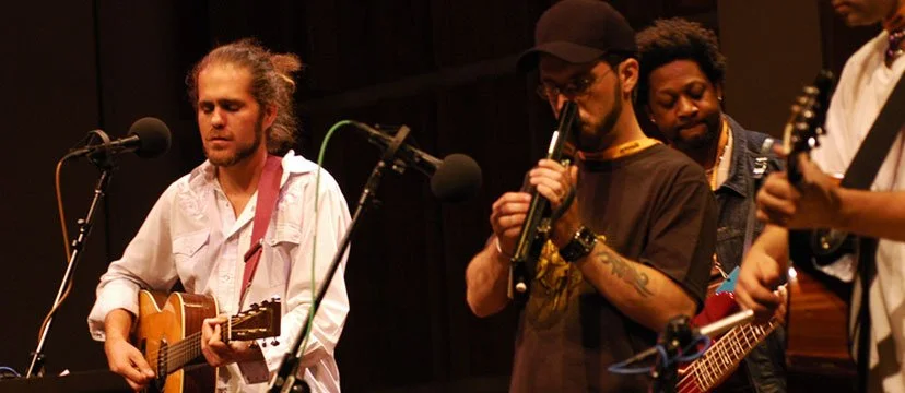 Four musicians performing on stage, three men and one woman, with guitars and a wind instrument.