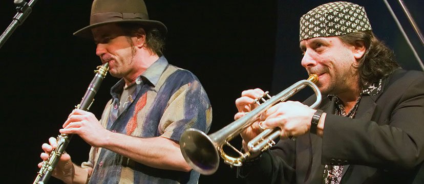 Two musicians playing wind instruments on stage, one with a hat and the other with a patterned bandana.