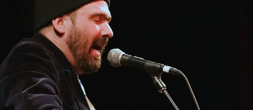 A man singing into a microphone on stage, wearing a black beanie and jacket.