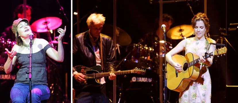 Three musicians performing on stage: a woman singing into a microphone with a hat and casual clothes, a man playing electric guitar with blonde hair, and a woman playing acoustic guitar in a floral dress.