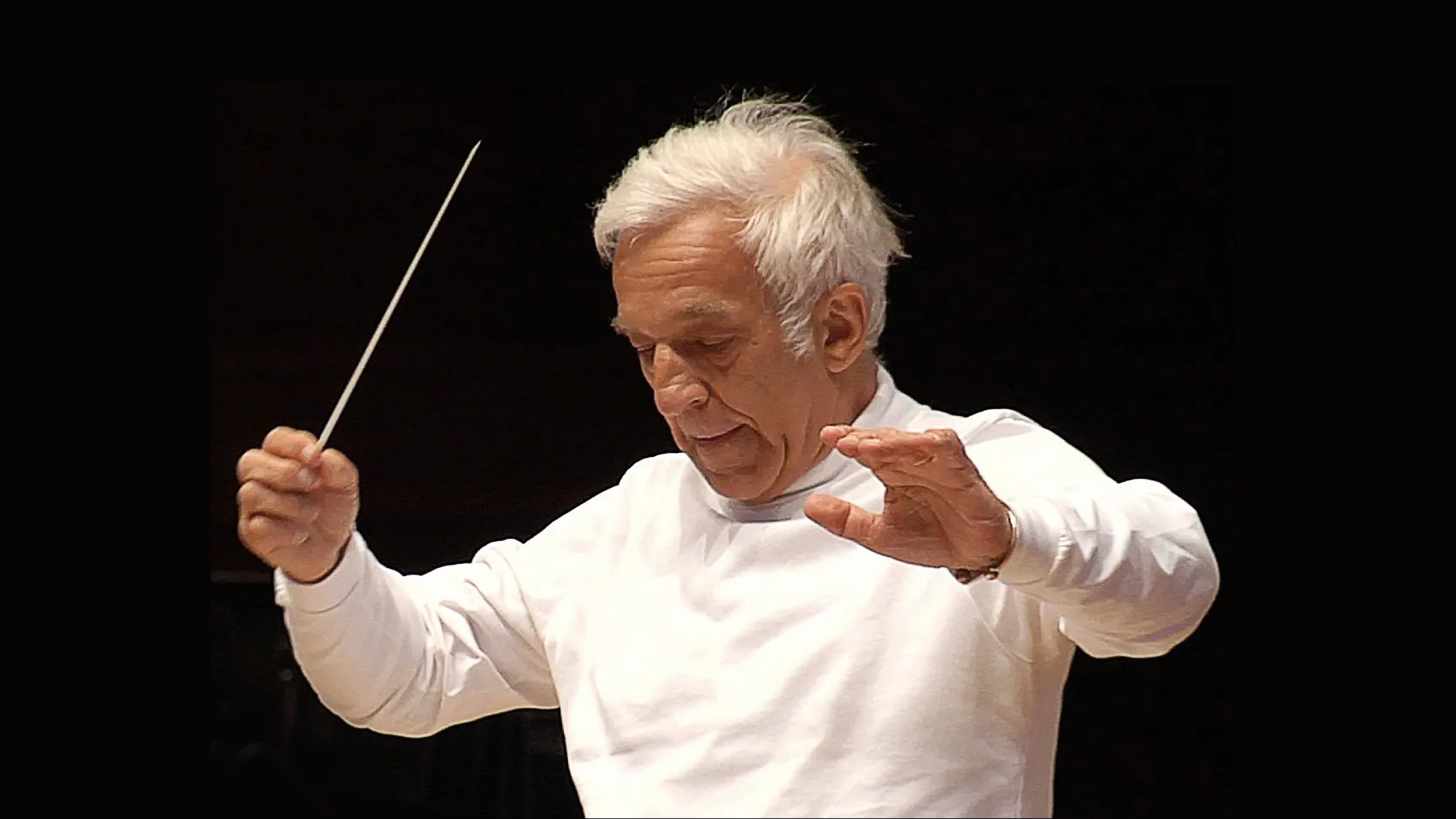 An elderly man with white hair conducting an orchestra, holding a baton in his right hand, with a focused expression, wearing a white long-sleeve shirt against a dark background.