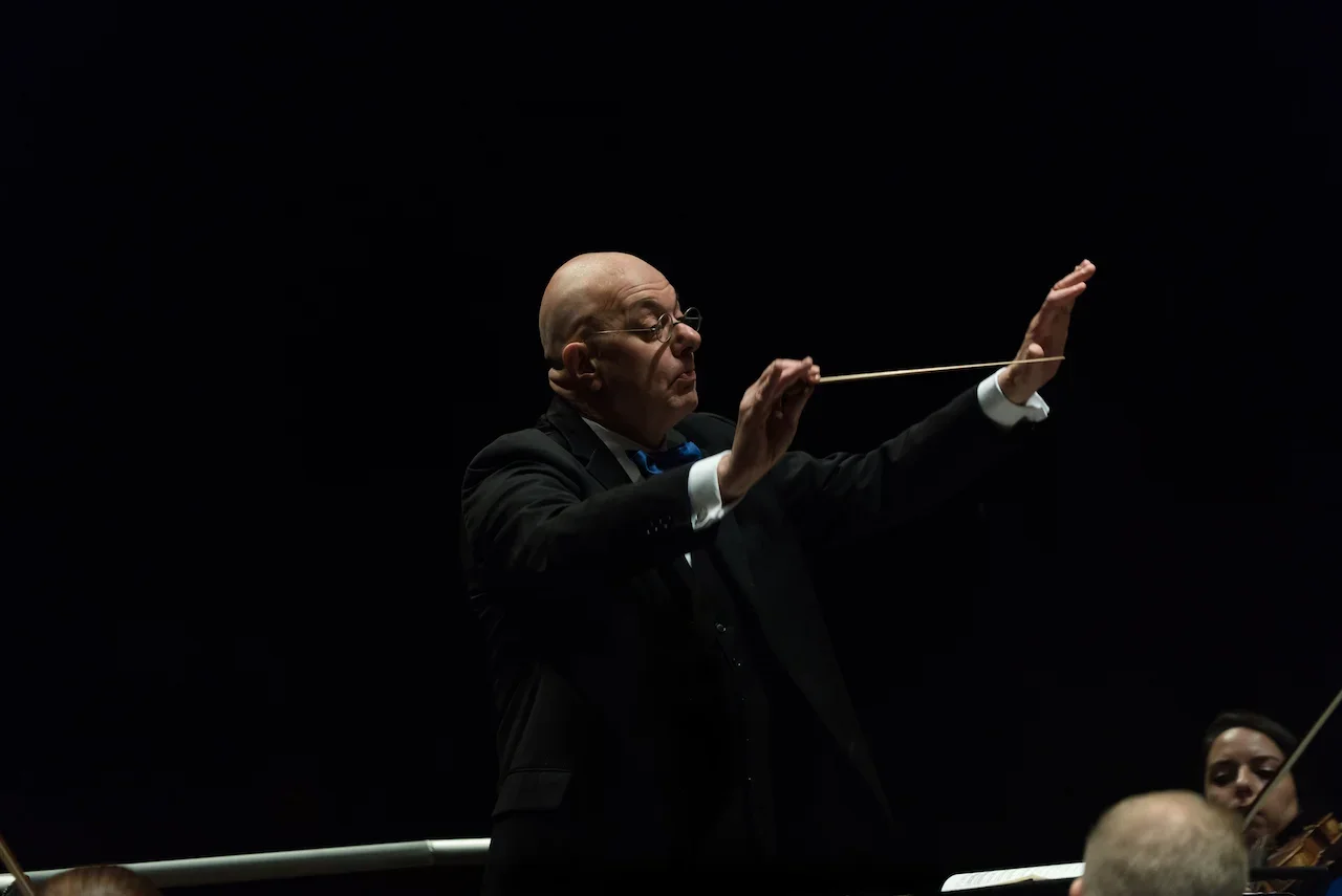 A bald man in glasses and a black tuxedo conducting an orchestra with a baton, with a dark background.