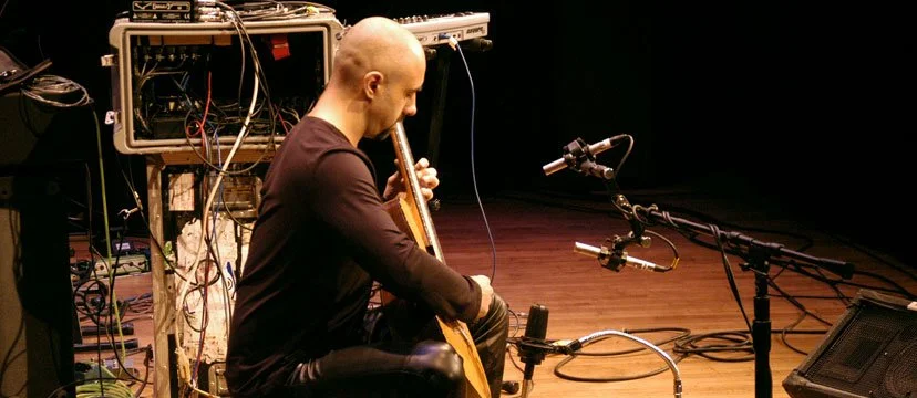 A man sitting on a chair playing a guitar in a recording studio with audio equipment and cables around him.