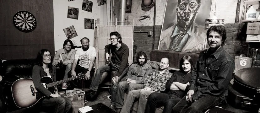 Group of seven people in a room, some sitting and some standing, with a guitar, paintings, photographs, and a dartboard on the walls.