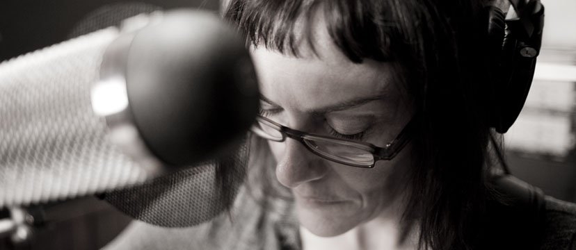 A woman wearing glasses and headphones recording vocals in a sound studio.