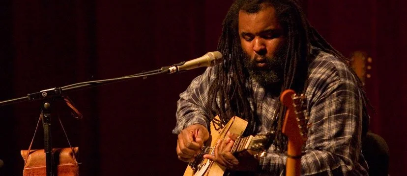 A man with dreadlocks playing an acoustic guitar in a dimly lit setting.